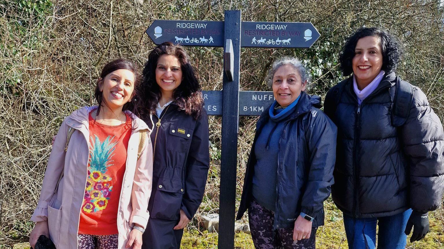 Geeta pictured with three other femal walkers, standing either side of a footpath sign saying "Ridgeway" on the top two arrows (pointing left and right). Other arrows below are concealed. There is a thick hedgerow of brambles and trees behind them. They wear autumn/winter coats and all smiling at the camera. One woman is older, about 60 plus, while the others appear to be in their 30s-50s.