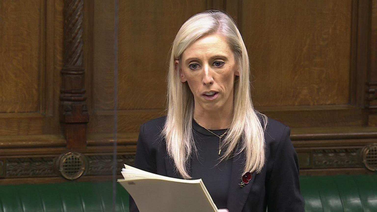 Carla Lockhart speaking in the House of Commons. She has long blonde hair, wearing a silver necklace, black blazer and black top with poppy pin on her lapel. She is holding pages. Wood panelling and a green leather bench is in the background.