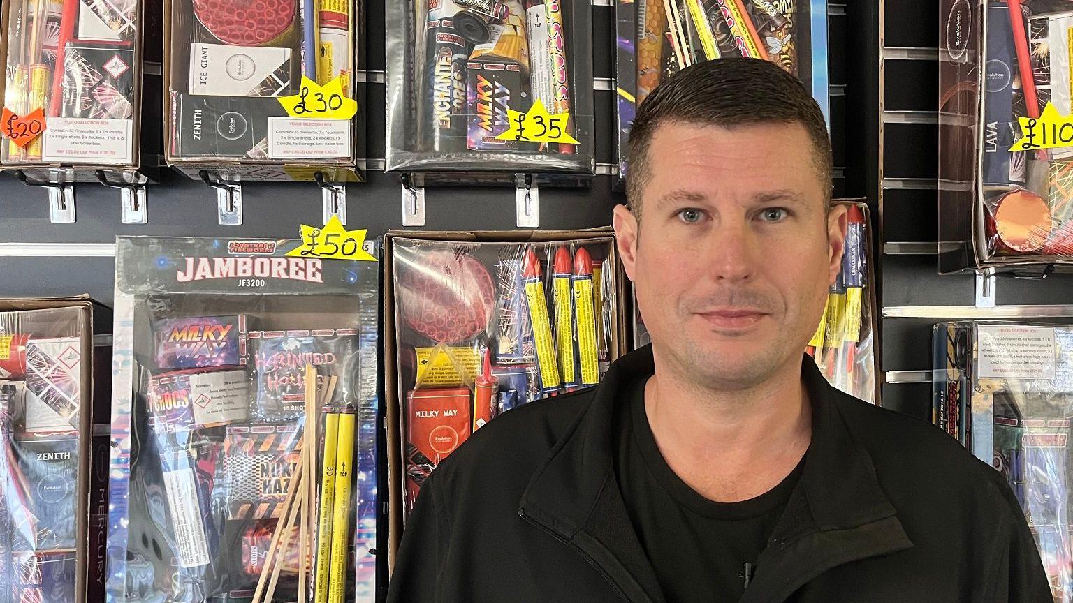 Mike Carter stands in front of colourful boxes of fireworks, which cover a wall of his shop. Yellow, star-shaped stickers advertise the prices. He has short, dark brown hair and wears a black T-shirt and a black jacket.