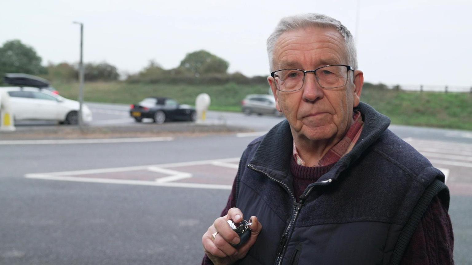 A man - with white hair and wearing glasses, a red shirt and jumper and a blue gilet - looks at the camera. He has a clicker-counter in his hand. Behind him cars are queuing at a junction.