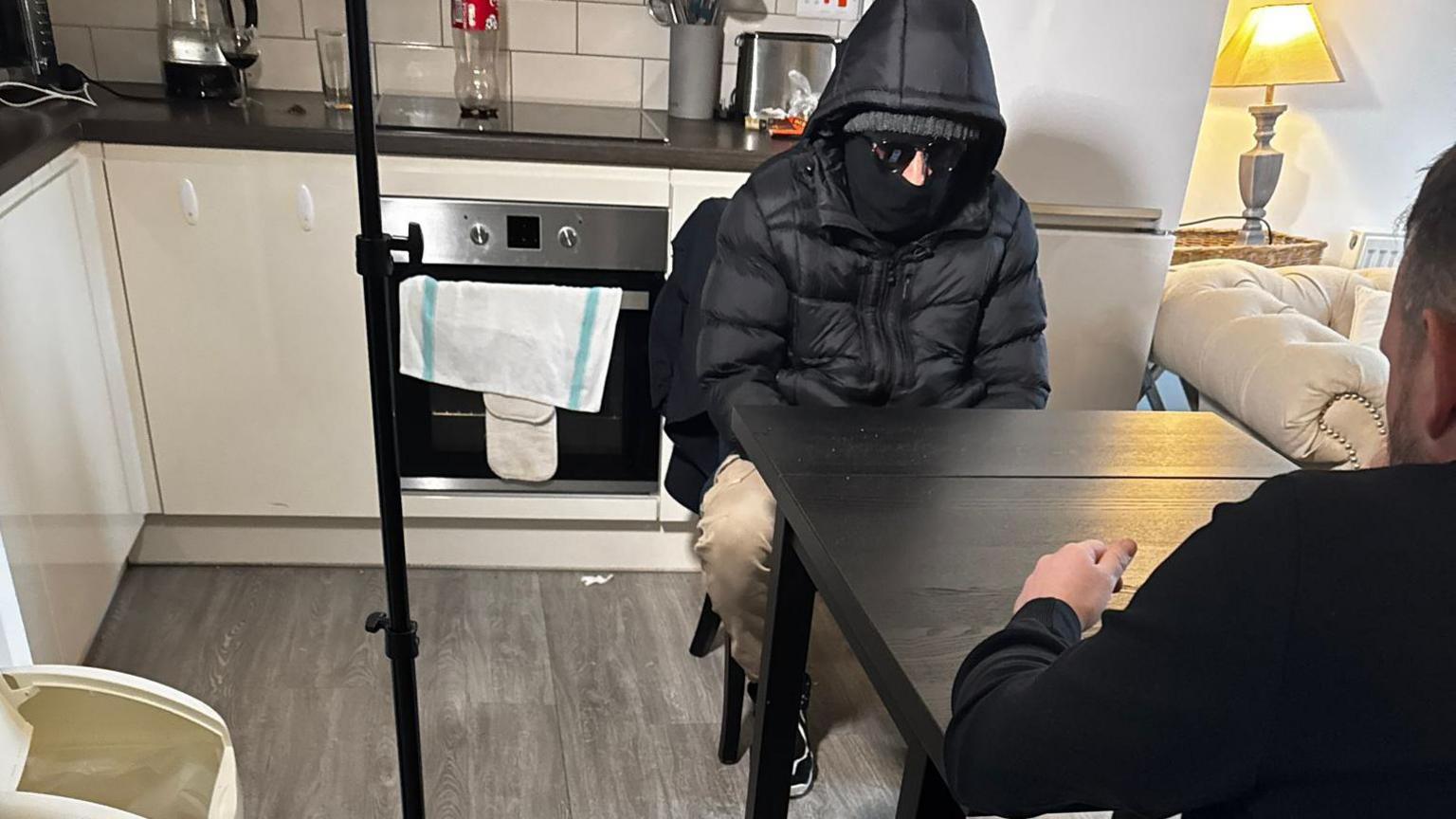 Zee, in a puffer jacket with its hood up and wearing sunglasses, sits at a table talking to a BBC journalist who is sitting opposite him.