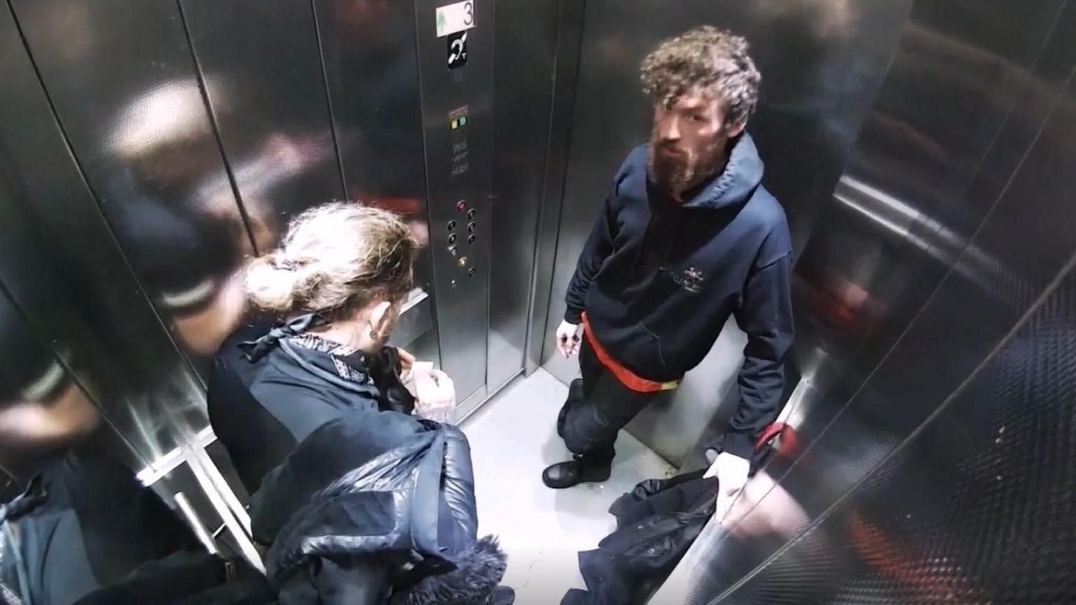 Two men wearing dark clothing are pictured in a lift. The men are seen from above. 