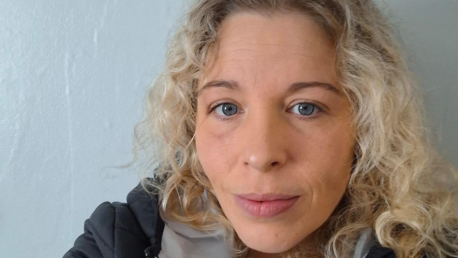 A woman with long curly blonde hair and wearing a black coat takes a selfie while standing against a pale blue wall.