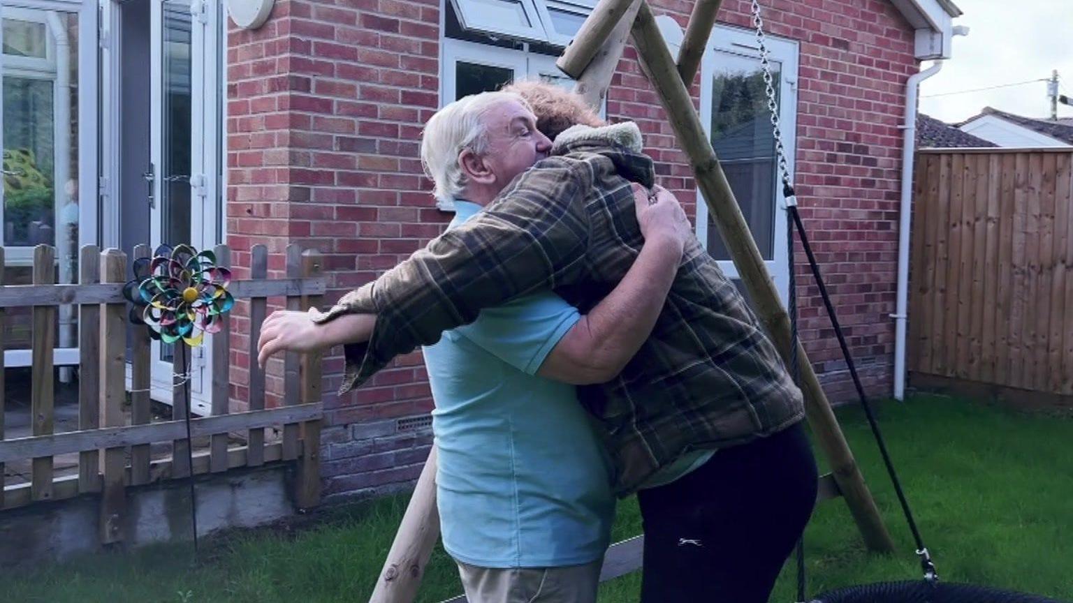 Christopher in a checked shirt hugging his father, Charles, a white-haired man in a blue -shirt, in the garden of a brick bungalow with a swing next to them