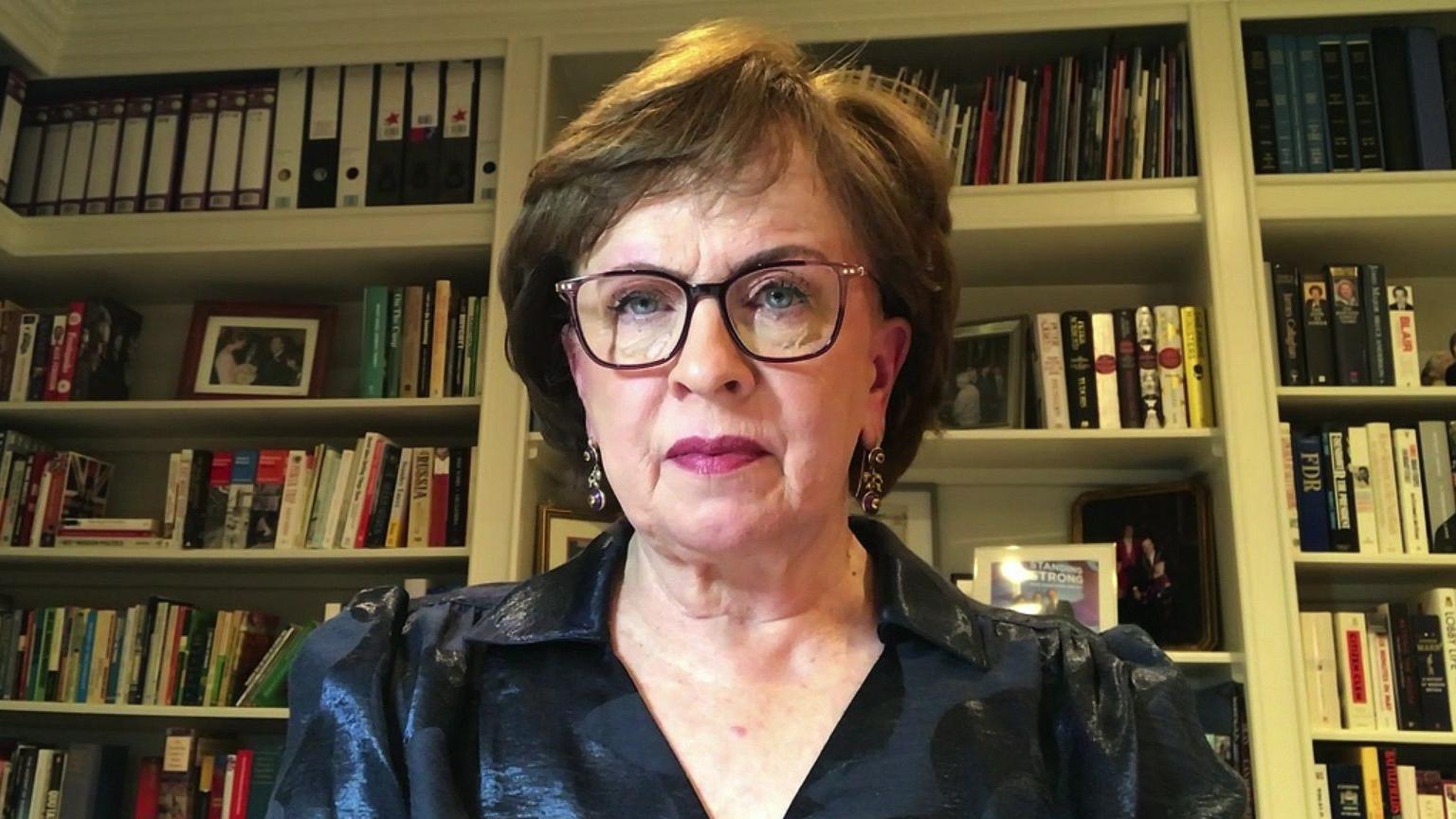 Diane Dodds sits in a room with books behind her. She has short brown hair, glasses and a satin navy blouse. 