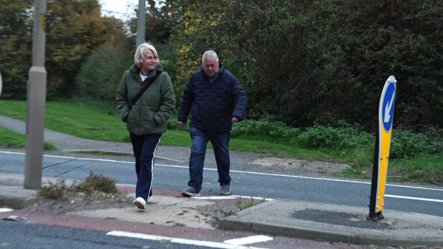 A woman and man hurriedly cross a busy A-road at dusk.