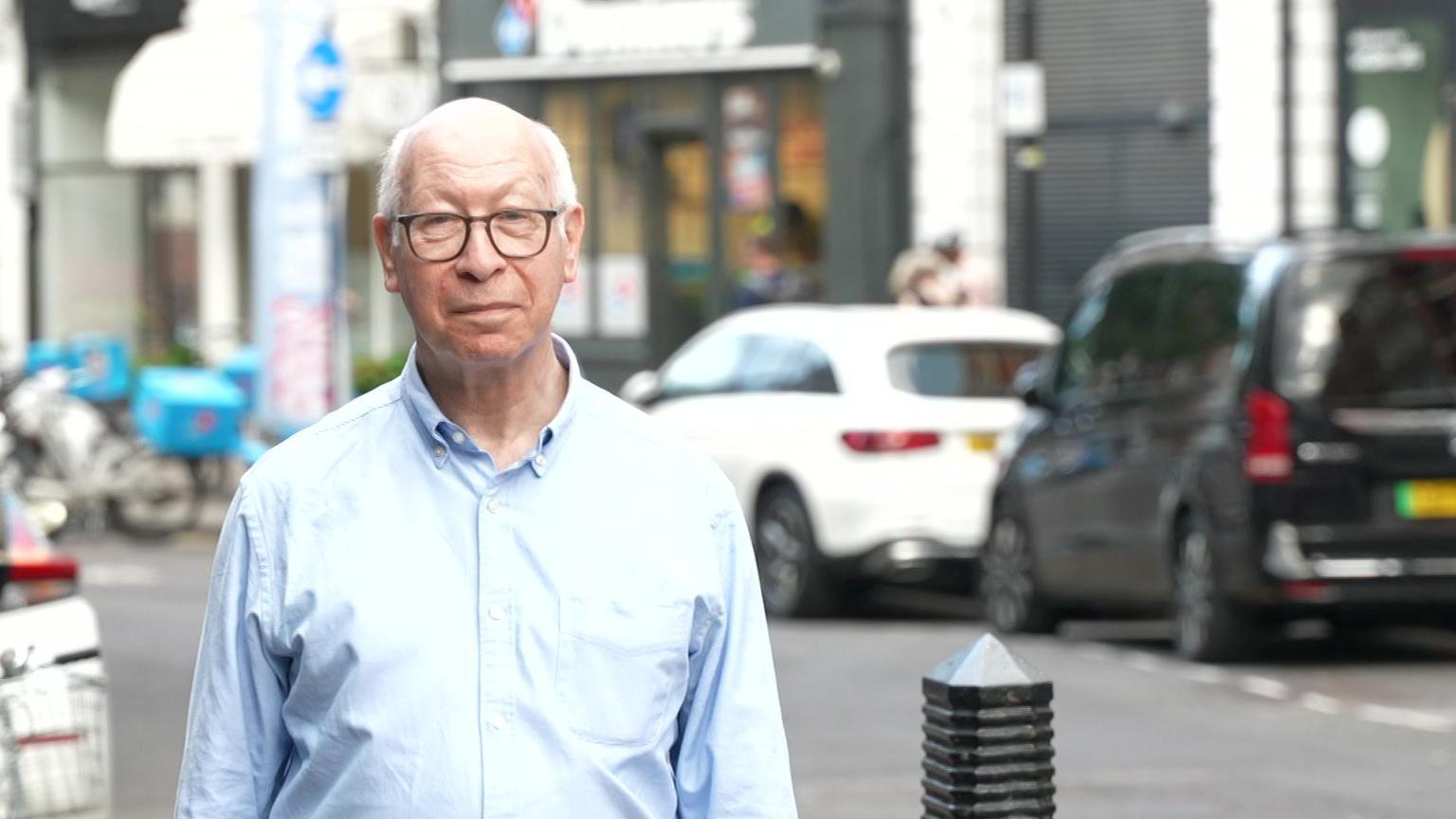 Gerald Oppenheim in a pale blue shirt standing on a London street. Blurred behind him are parked cars. 