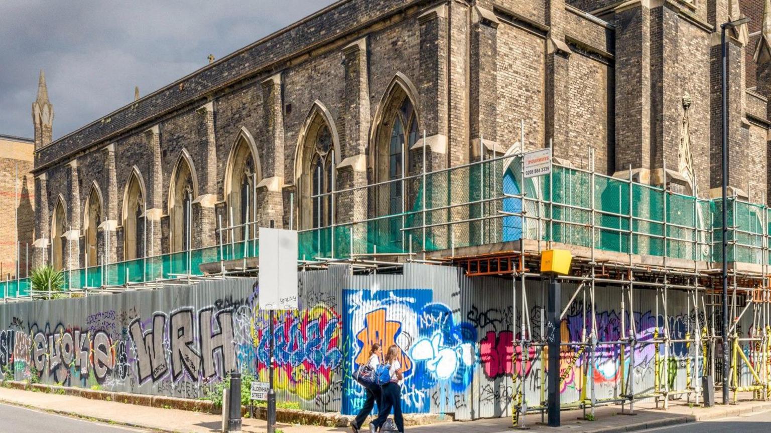 Exterior of a Gothic-style stone church with scaffolding and colourful graffiti-covered hoarding, under cloudy skies.