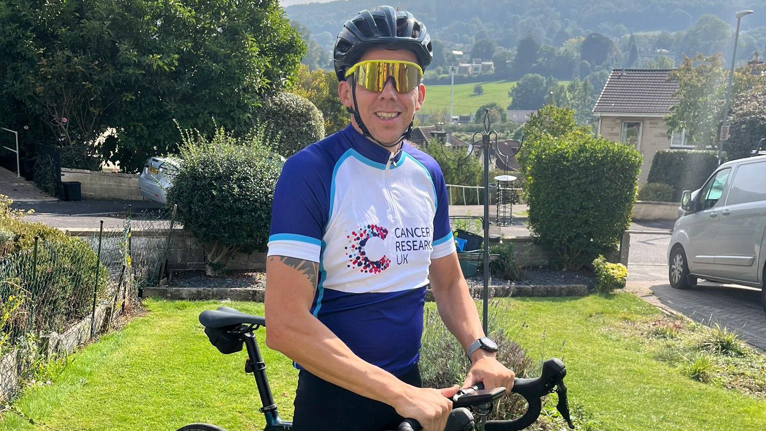 Mark Parsons wearing a blue and white Cancer Research UK T-shirt. He is pictured with his bike and is wearing a helmet and cycling sunglasses