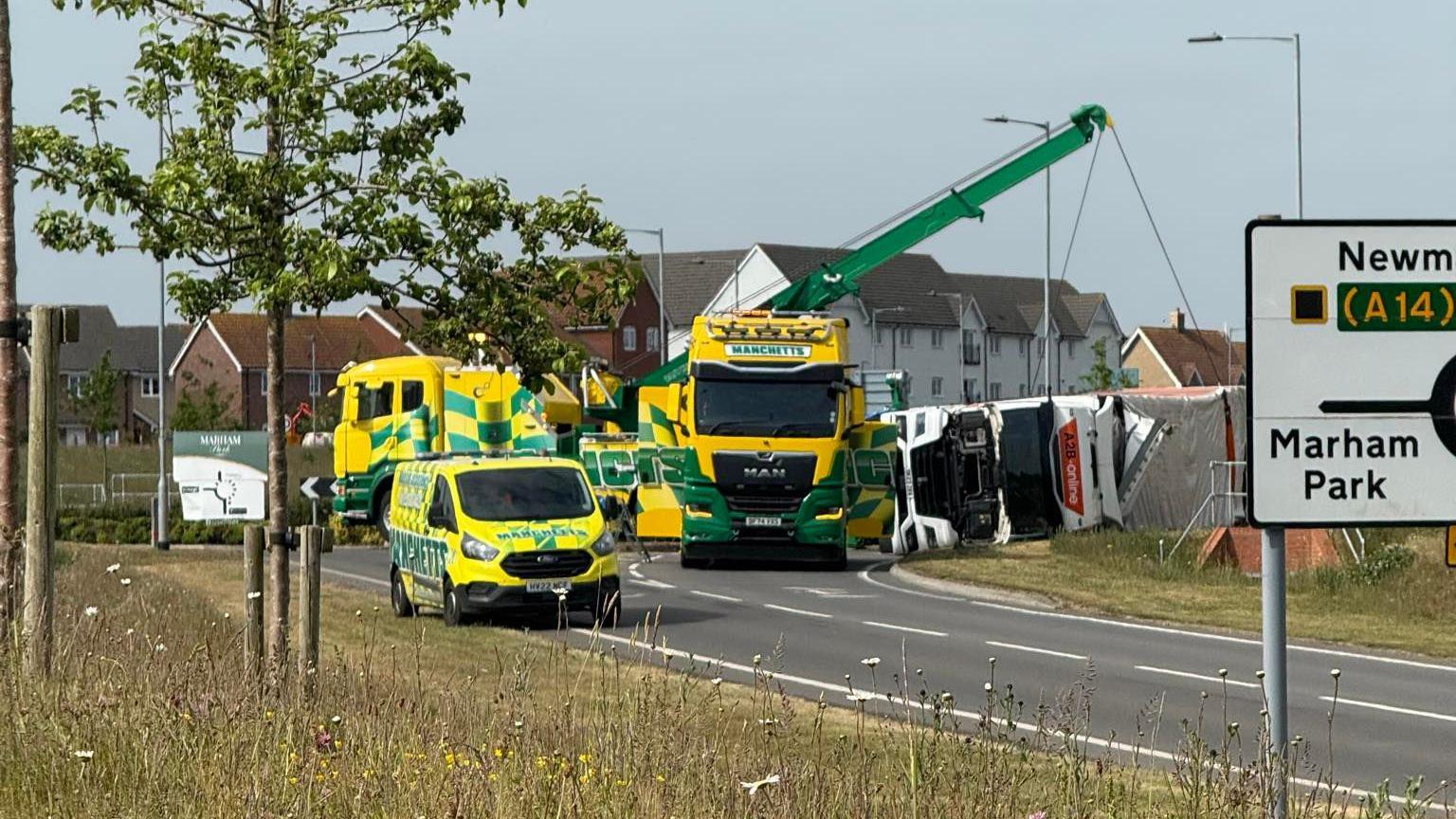 A specialist lorry with a crane on it lifts the overturned lorry off the road. Other recovery vehicles are parked nearby.