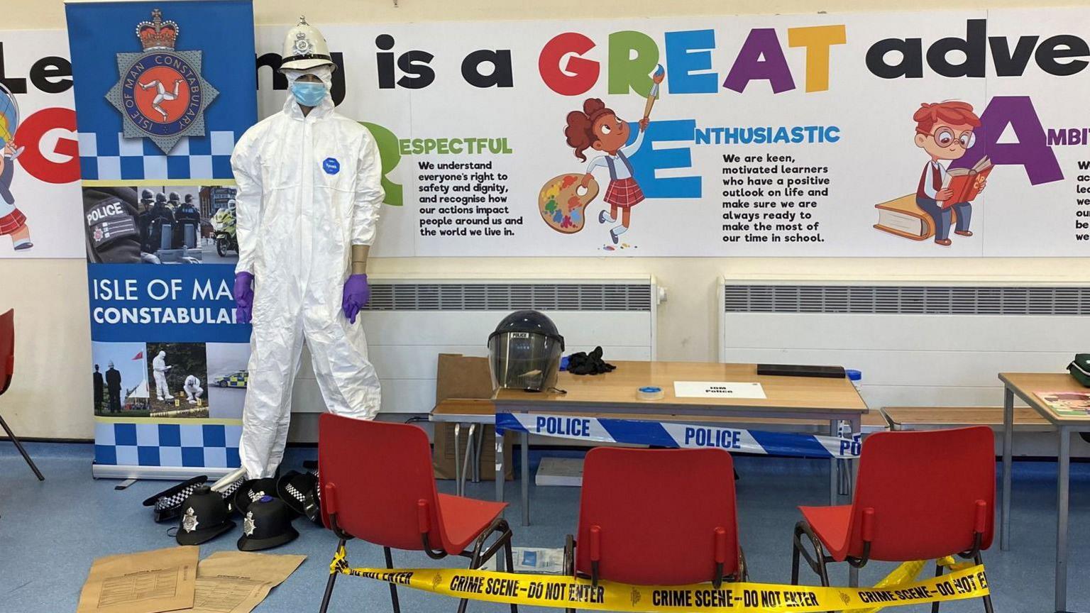 The CPR mannequin is "standing" upright on the left as part of a display in a primary school classroom. There is a table with three chairs covered in police tape, and a helmet on the table.