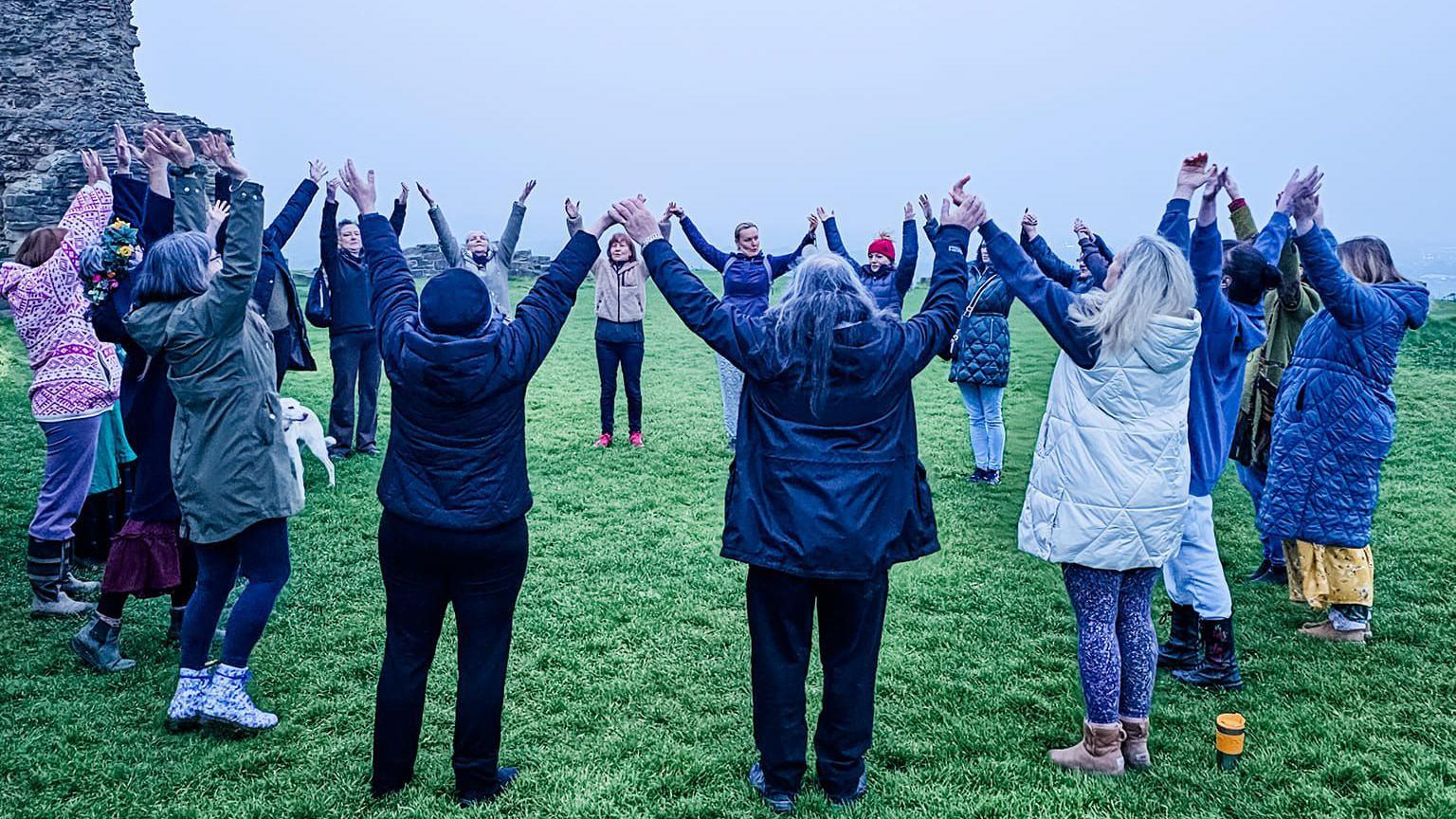 Seventeen people stand in a circle with their arms raised holding hands on a grey morning with castle ruins in the distance.