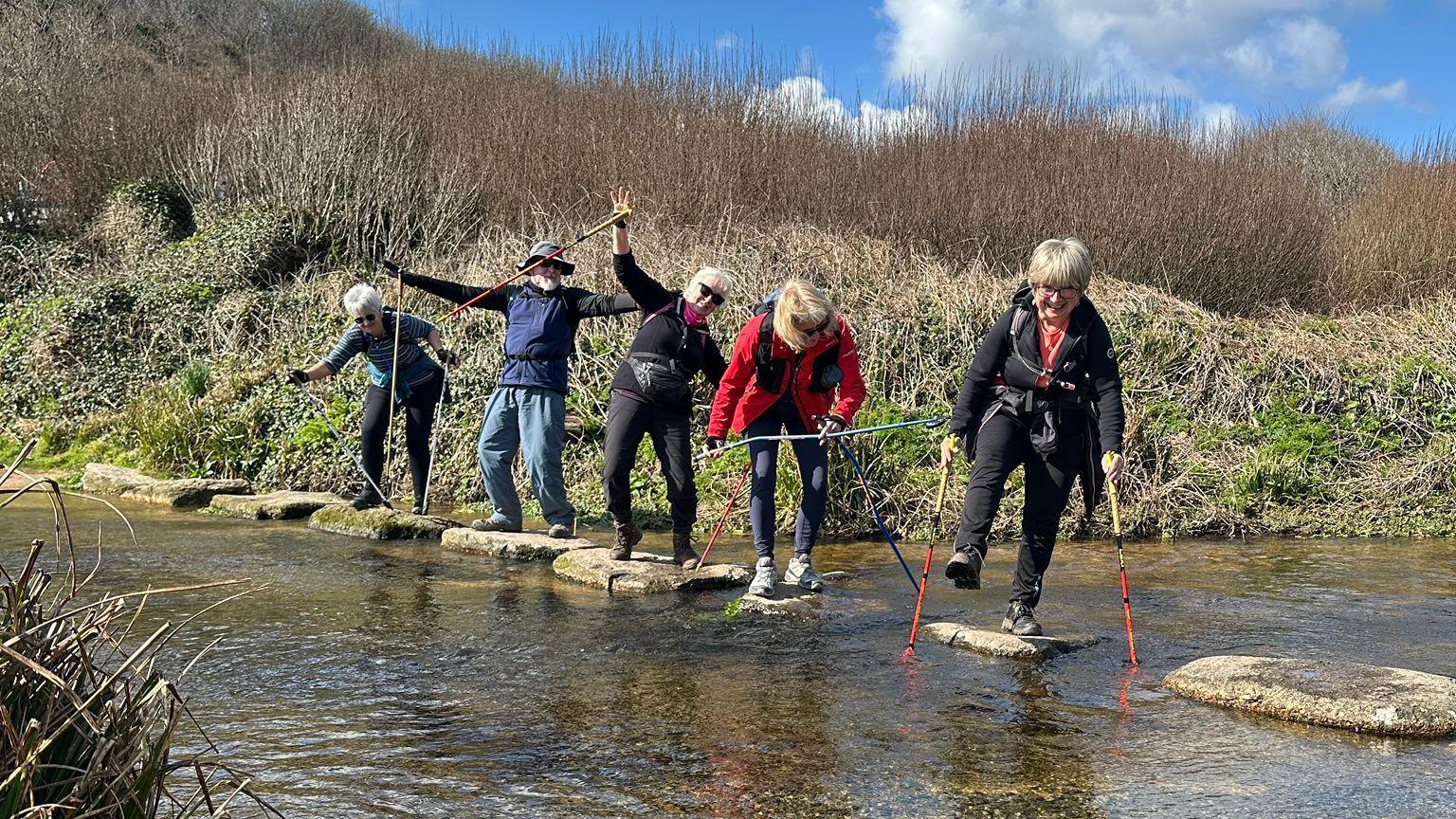 Four women and a man from Walk Kernow pose for a fun picture on stepping stones over a waterway near Land's End in Cornwall. They are all pretending that they're about to fall into the water. The woman at the front of the group is standing on one leg.