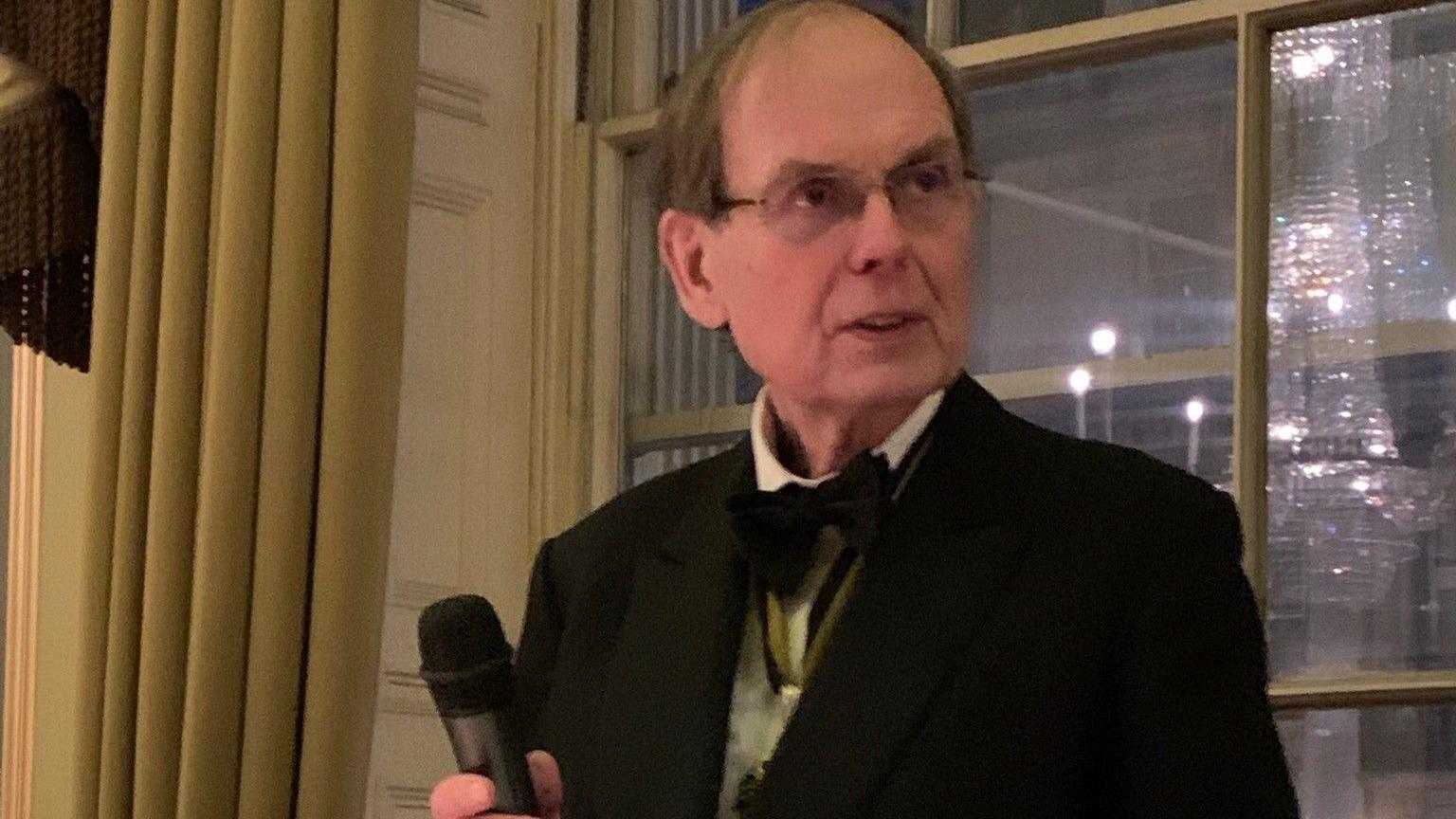 A balding man with glasses wearing a black tuxedo and holding a microphone, with a window behind him.