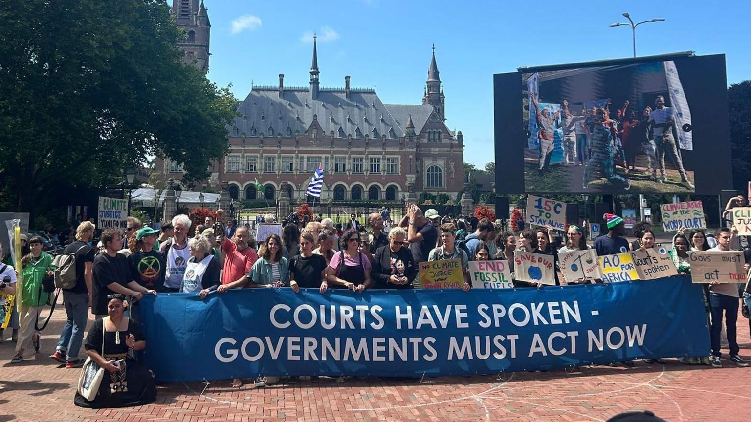 A group of roughly 50 people stand outside the Peace Palace building in the Hague. They are holding a blue banner with white letters that read "Courts have spoken. Many are holding placards calling for action on climate change Governments must act now". In the background is a large screen with more activists.