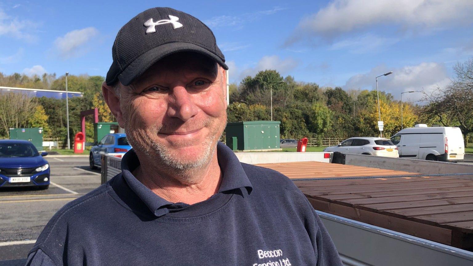 Malcolm Johnson is wearing a blue sweatshirt and polo shirt.  He is smiling at the camera and has a black baseball cap on