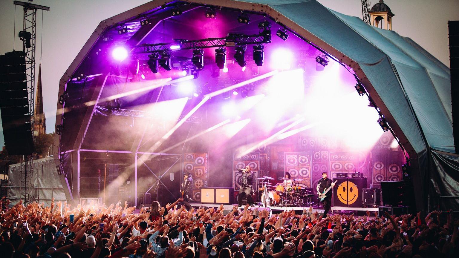 Skindred performing at Bristol Sounds. A large crowd of people can be seen with their arms up. The band is on stage with instruments including drums and guitars. 