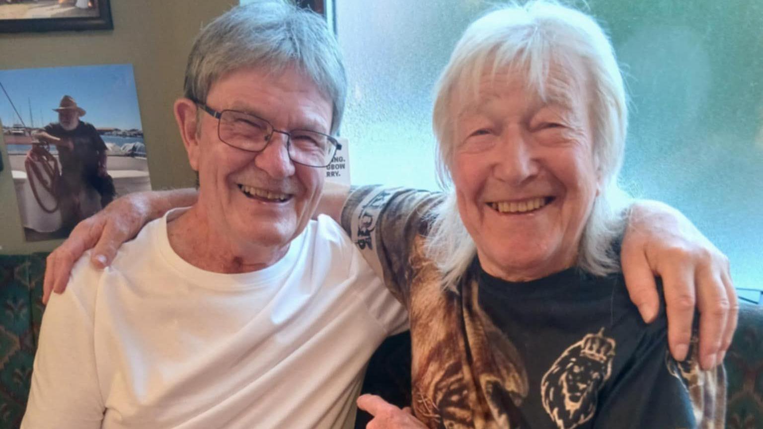 Chris and Snip together in 2025. They sit with arms around each other smiling broadly. Chris wears rectangular metal glasses and a white T-shirt, Snip has a fringe and shoulder length white hair, and wears a black t-shirt with lions on.