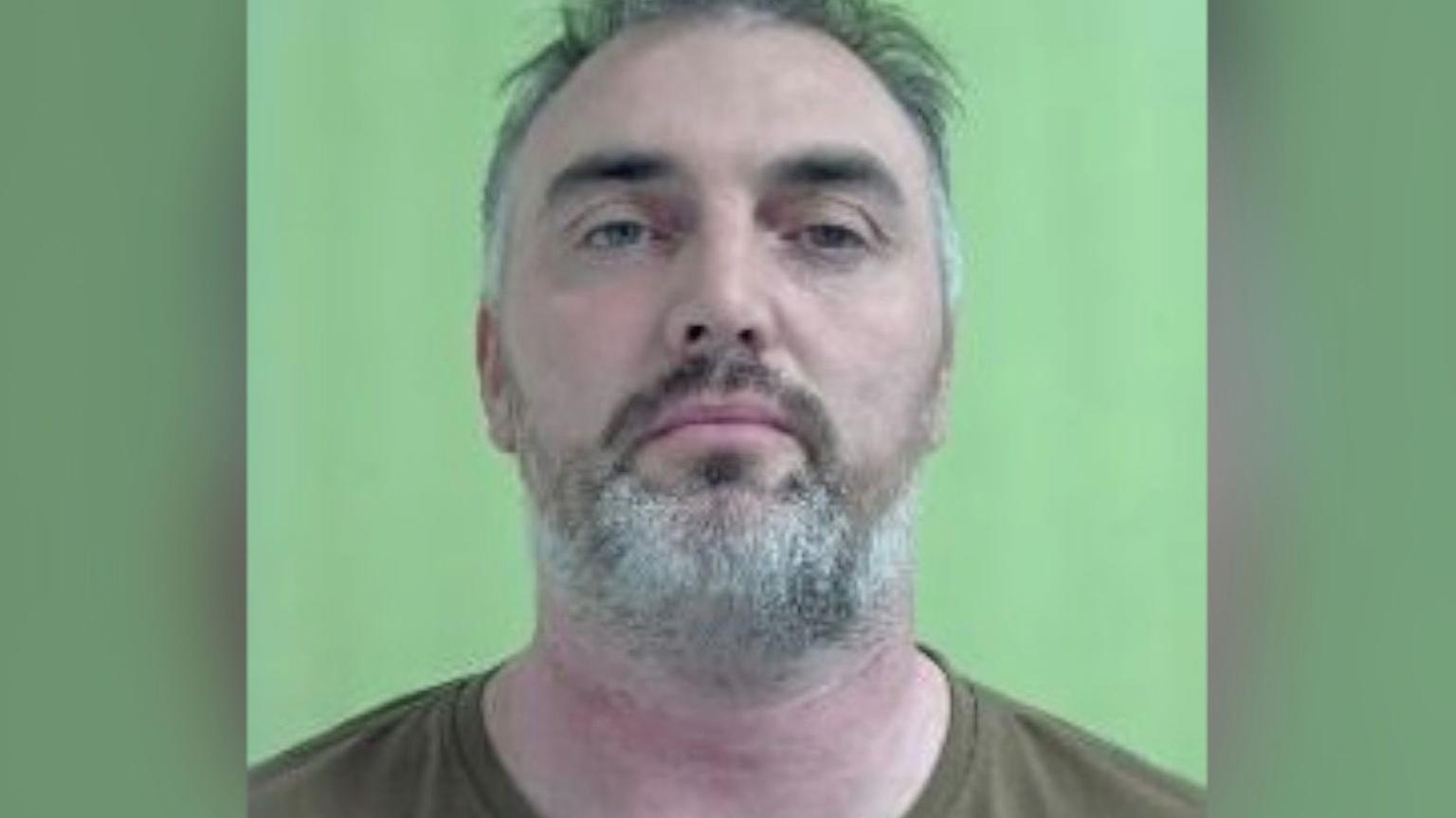 A mugshot of John Skillen a man with grey hair and a grey beard with a green background.