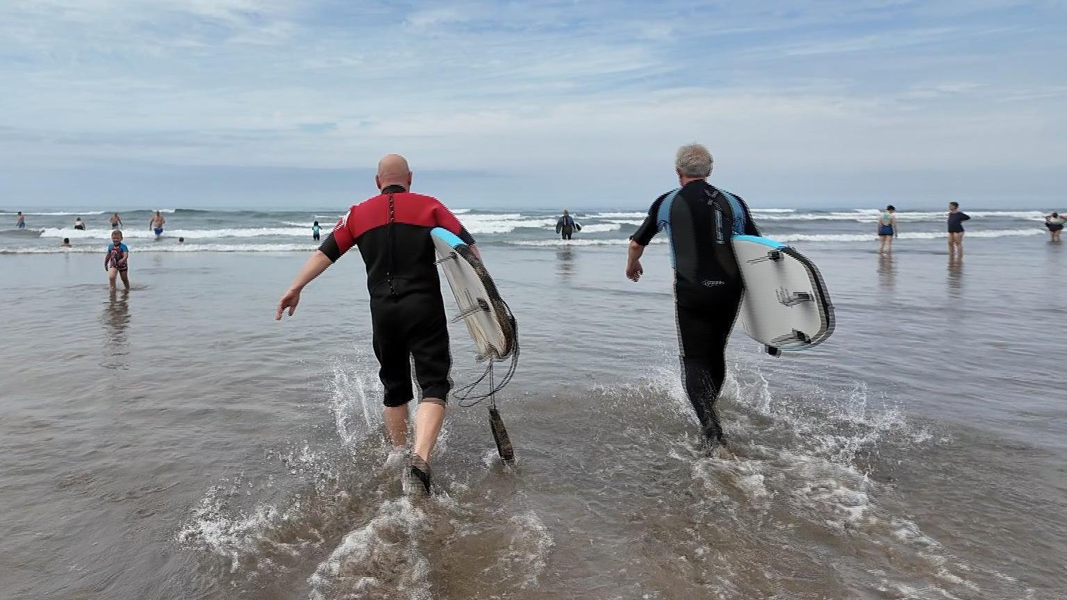 Two men in wetsuits are walking into the sea at Bude, Cornwall. they are both holding surf boards and 
