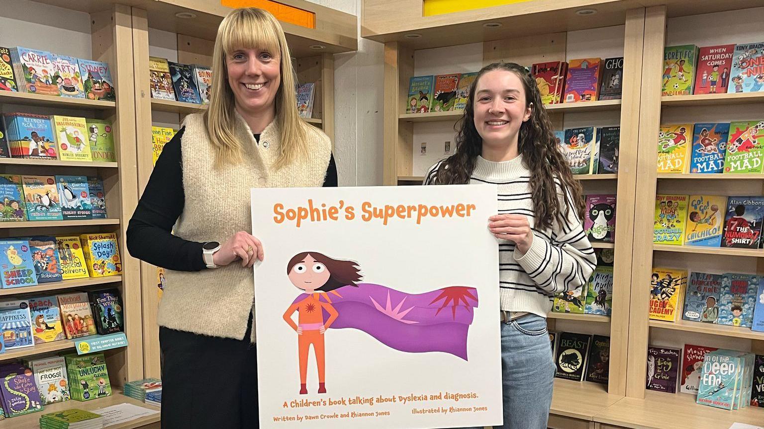 Dawn Crowle and Rhiannon Jones. Dawn has long blonde hair and is wearing black with a cream knitted vest. Rhiannon has long dark hair and is wearing blue jeans and a striped jumper. They are stood side by side, holding a large copy of the Sophie's Superpower book cover. It is white with an illustrated superhero wearing an orange suit with a purple cape.