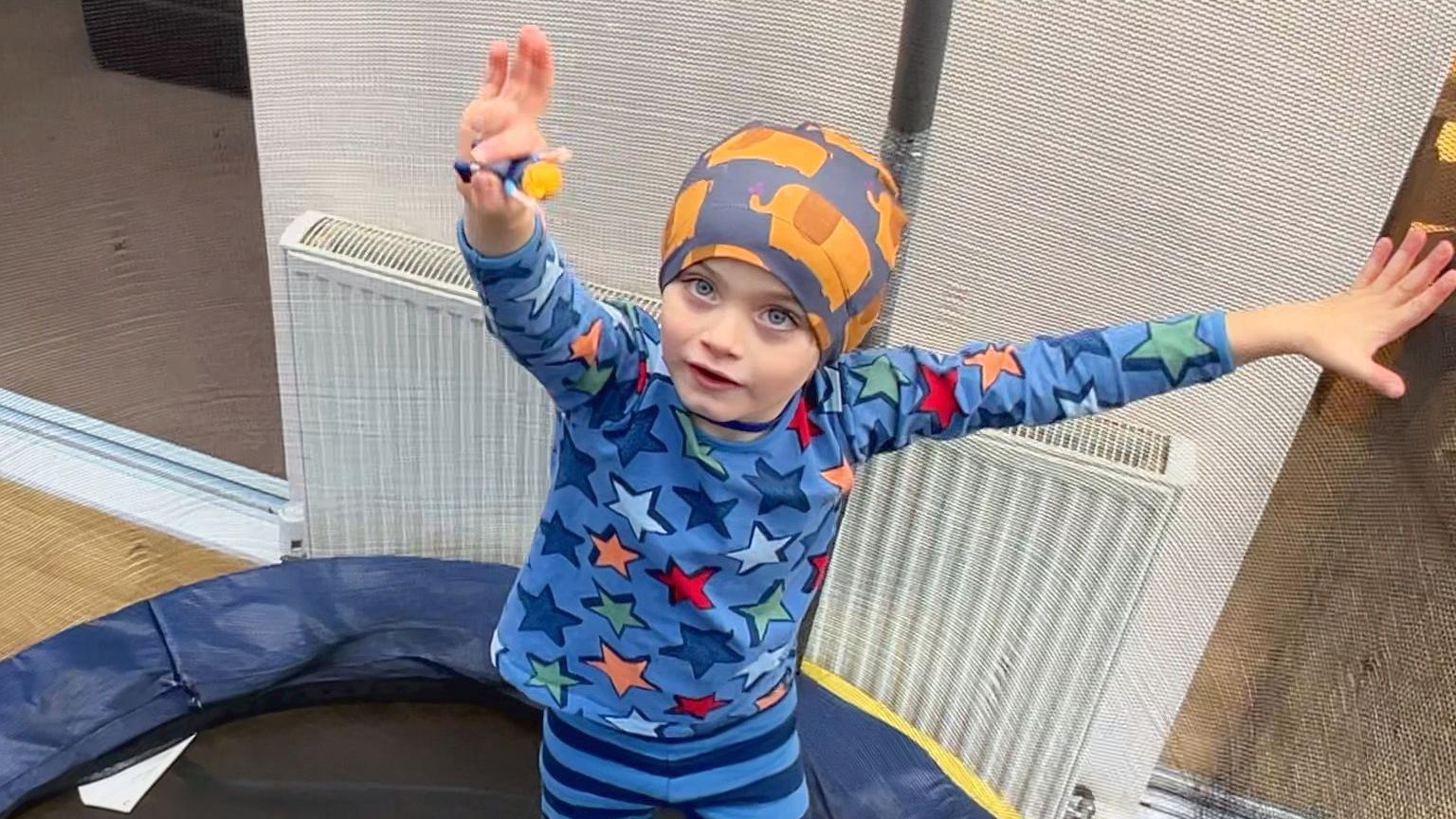 Lochlin wears a fabric hat, and is holding a toy. He wears a bright blue top with colourful stars, and stripey trousers. He's waving on his trampoline. 