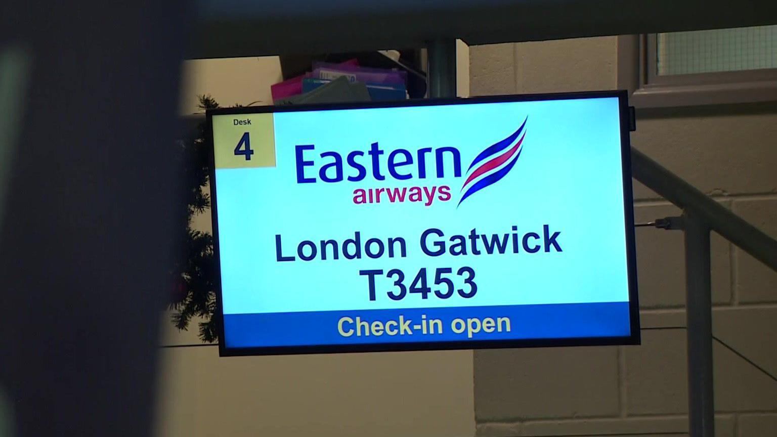 A screen inside an airport showing the check-in for an Eastern Airways flight to London Gatwick is open. The flight number is T3453 and the check-in is at desk four.