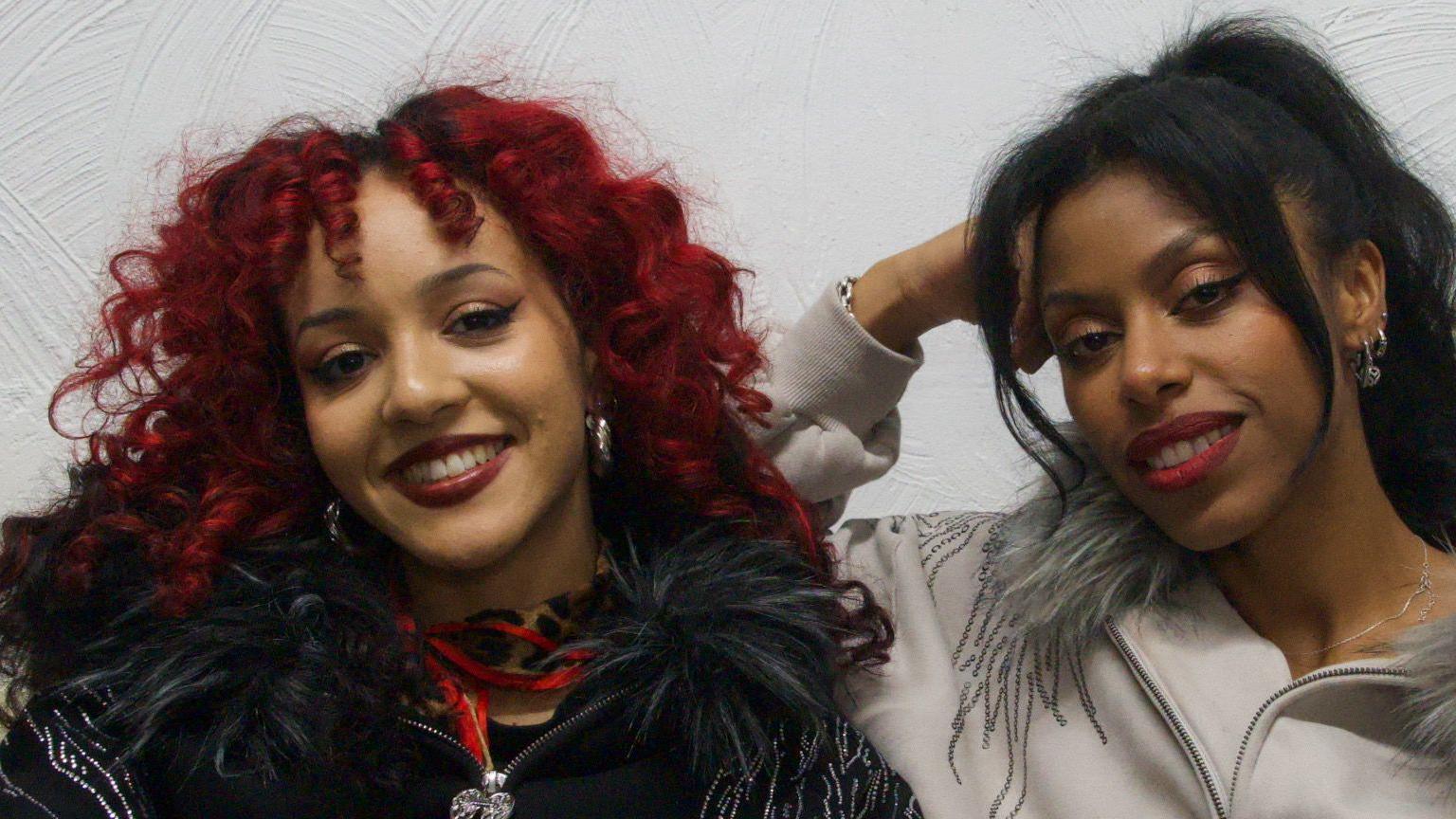 Nova Twins sit on a sofa looking at the camera. Georgia South is on the left side of frame. She has red curly hair and wears black clothing, with dark red lipstick. Amy Love sits on the right side of frame. She is wearing a grey 