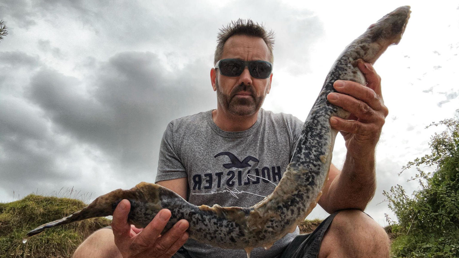 A man, wearing sunglasses, holding a lamprey. The lamprey looks like a long eel. 