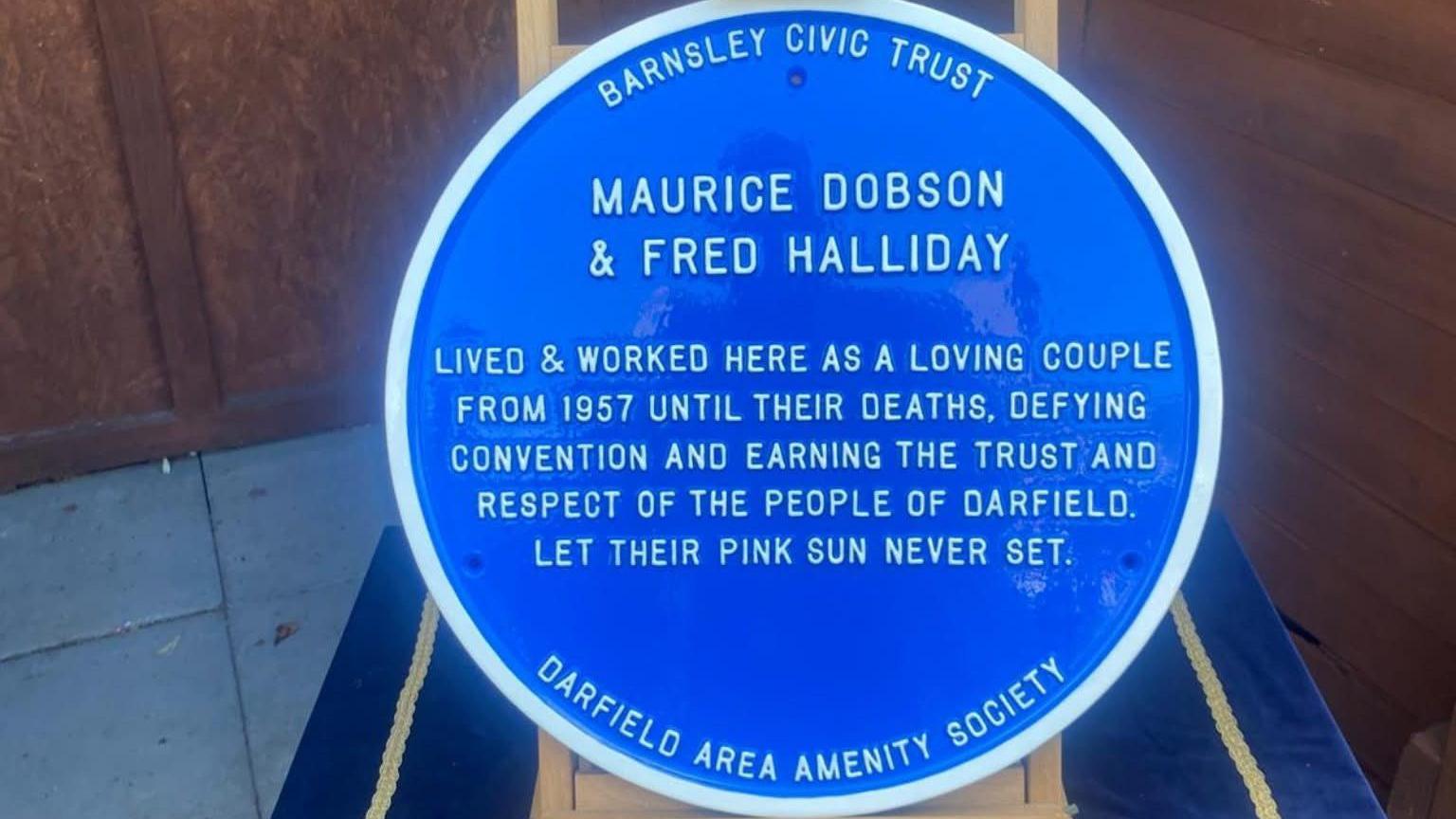 A blue plaque on a wooden stand. The plaque reads: "Maurice Dobson & Fred Halliday. Lived & worked here as a loving couple from 1957 until their deaths, defying convention and earning the trust and respect of the people of Darfield. Let their pink sun never set."