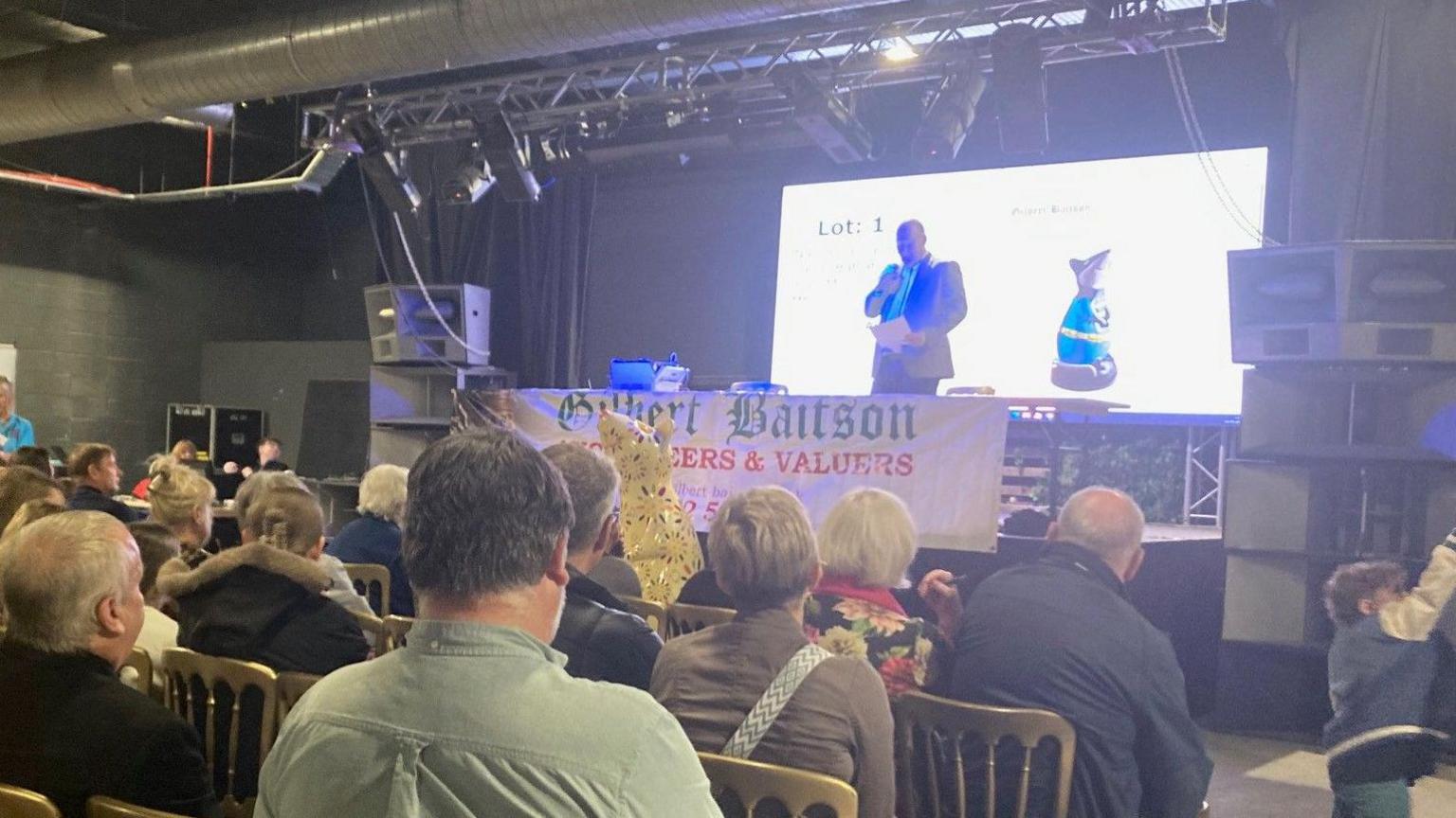 The auctioneer stands in front of a screen that displays one of the rat sculptures.  People are sat on chairs with their backs to the camera during the bidding.