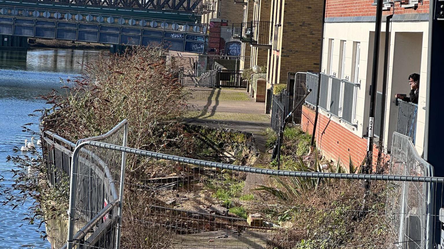 A fenced off footpath with a sinkhole in it- placed between the River Nene and a block of flats on the other side of it.