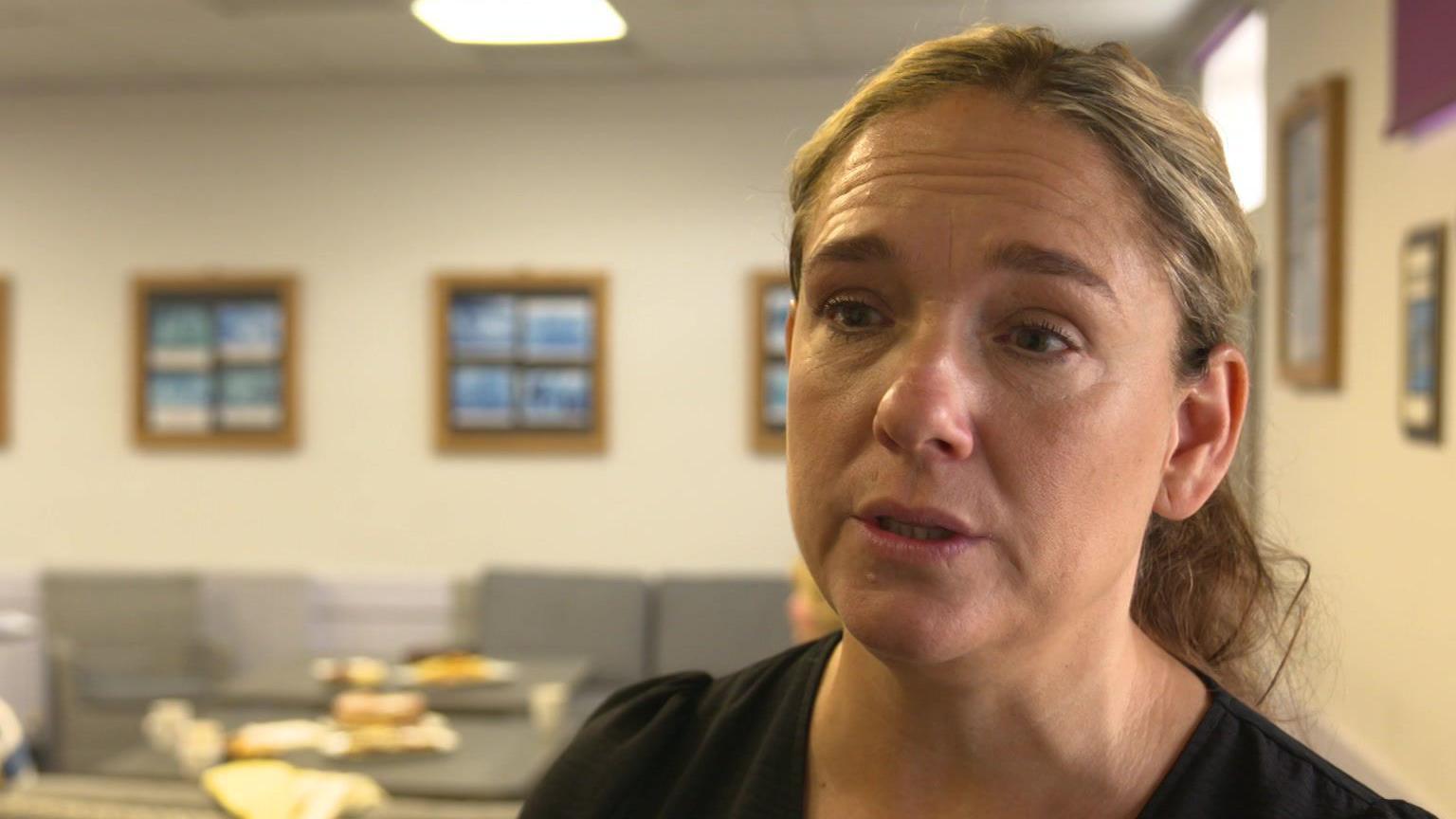 Ruth Abram is talking to the the interviewed in this candid image. She has her blonde hair tied back in a ponytail and is wearing a black top