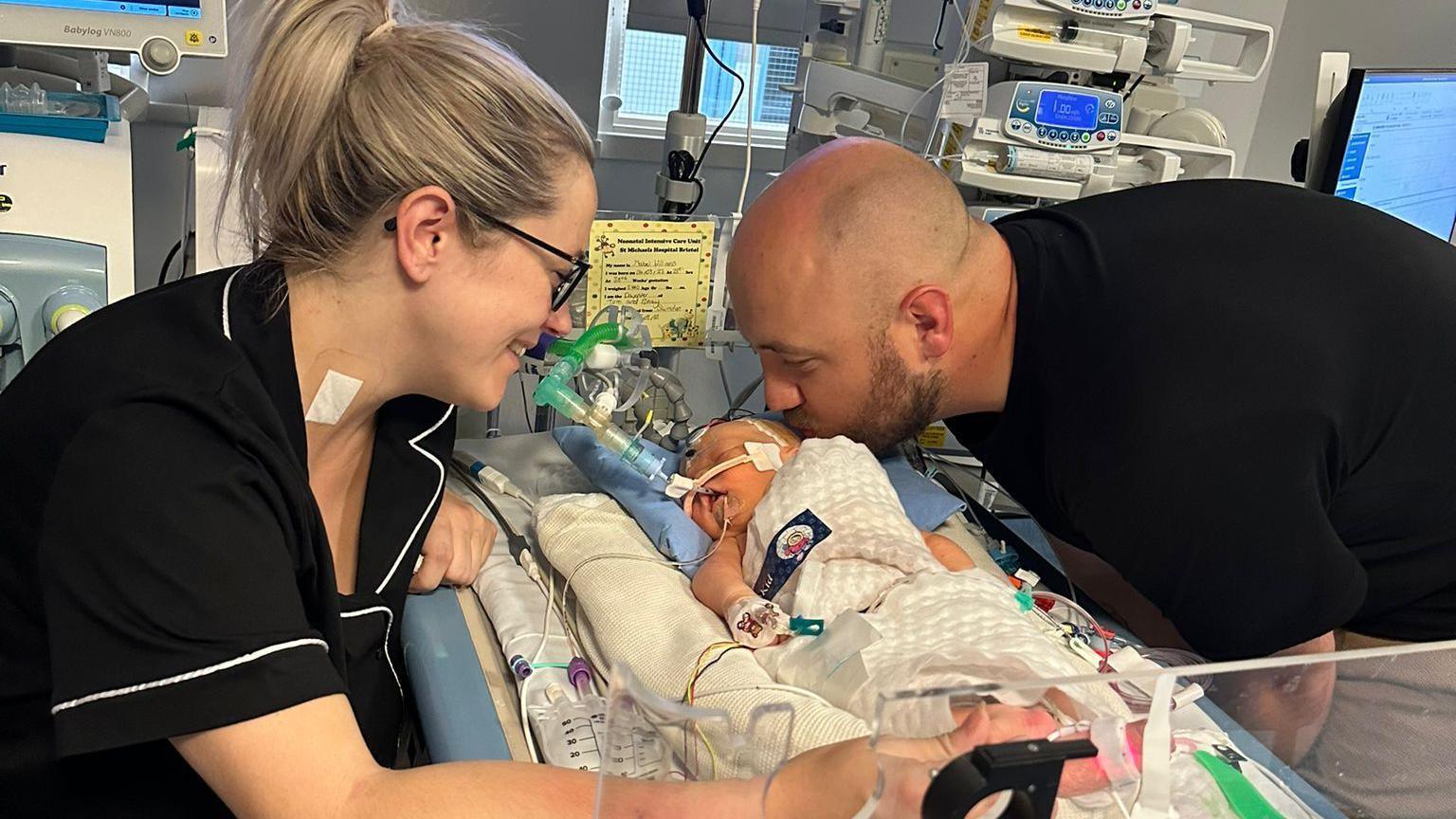 Tom and Becky Williams are perched over Mabel who is surrounded by wires & machines in a hospital. Mabel is lying down on a bed. Tom is kissing her on the head while Becky smiles.