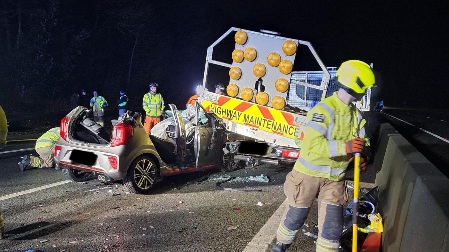 A photo of the scene of the collision on the M11. Firefighters and other emergency service personnel can be seen working around the area. A car that is missing its roof, doors and boot can be seen at the back of a highways vehicle.