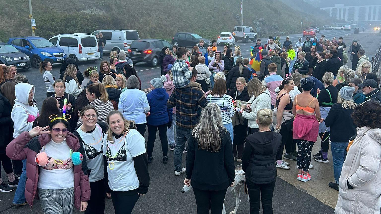 A large group of people gather outdoors in a foggy car park near a grassy hill. Many wear bras or bralettes over their normal clothing.