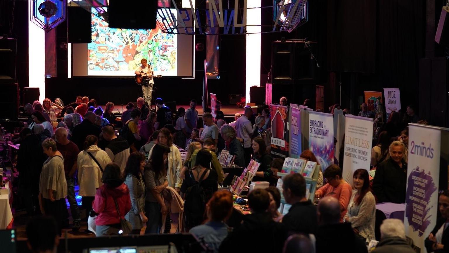 A large room is full of people milling around and looking at vendors selling books at stalls. A man is playing the guitar and singing on a stage in the background.