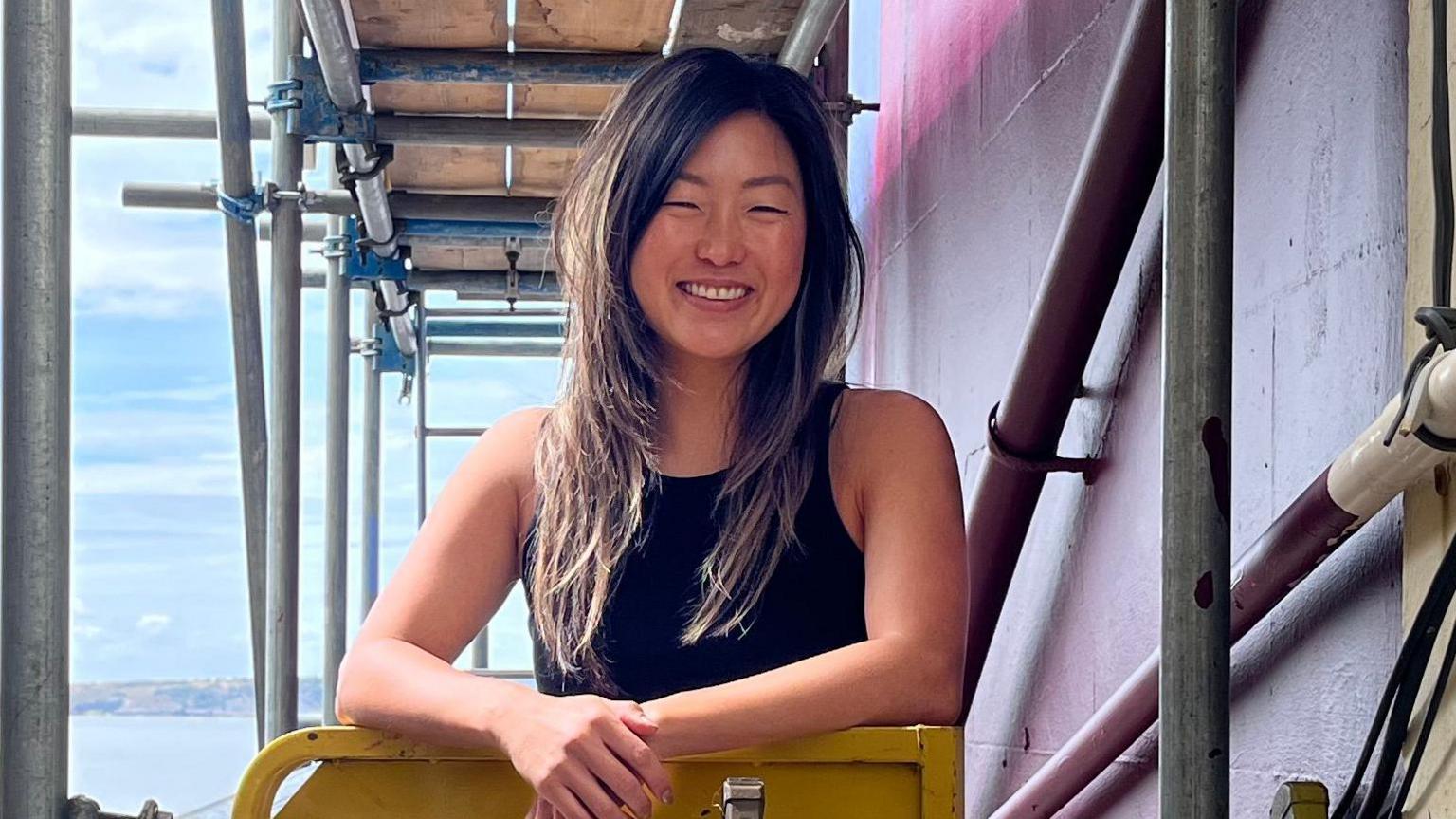 A woman standing on scaffolding smiling. She is stood next to the side of a seafront pub she is painting. Behind her in the distance is the Bristol Channel.