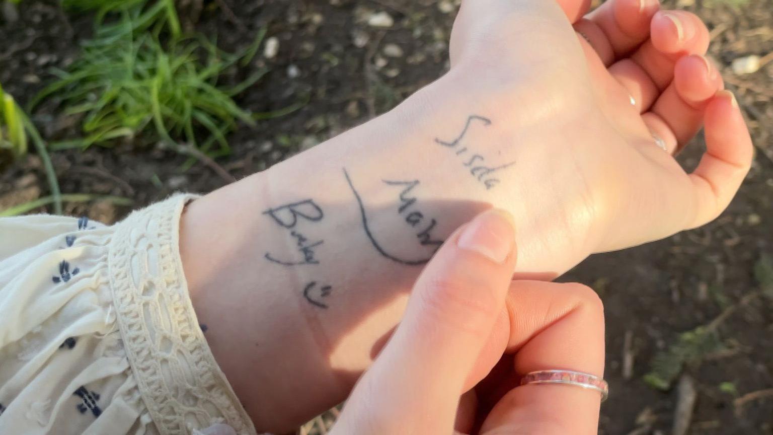 An image of Mary's left wrist showing the tattoo of Alex's handwriting. The photo is taken from above, there is grass underneath Mary's wrist in the background and she has a white floral blouse which has been pulled up her arm slightly to show her wrist. Her right thumb is positioned as if pointing towards the word 'Sisda' and she has a pink gemstone and silver band on her right index finger. Another part of the tattoo which is in vision reads 'Bucky' with a smiley face.