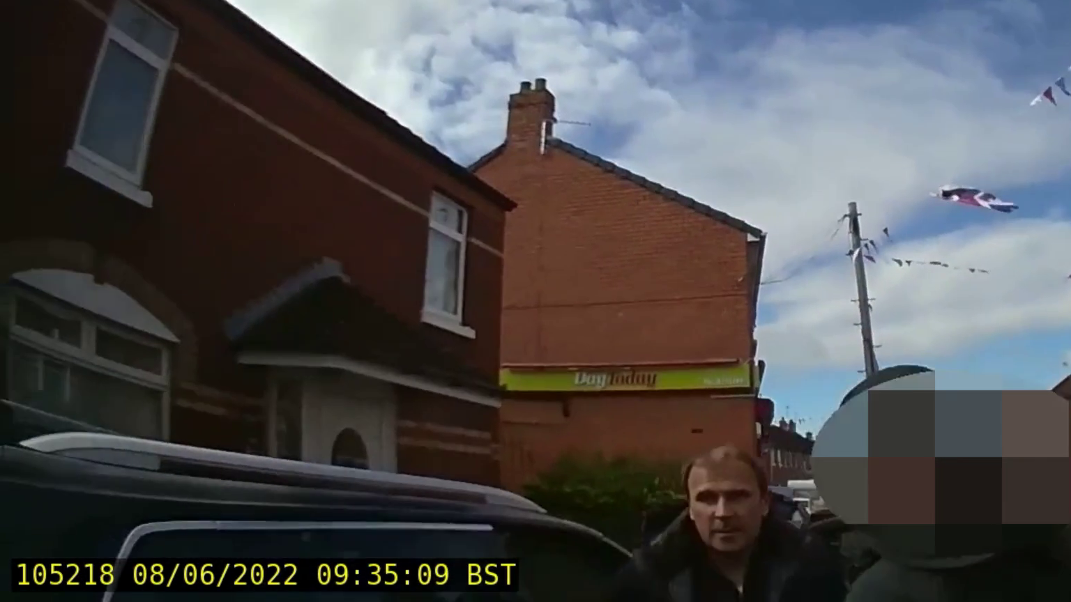 You can see two red brick buildings, one is a home, then other is a shop with Day Today signage on the side.
A union flag can be seen flying in the background.
In the foreground, a man - Winston Irvine, it stood beside a car.
Beside him is a police officer - the PSNI has blurred his identity.
On the bottom left hand side of the picture there is yellow text which gives the date the footage was taken - the 8th of June 2022 - and the time - 9:35 am