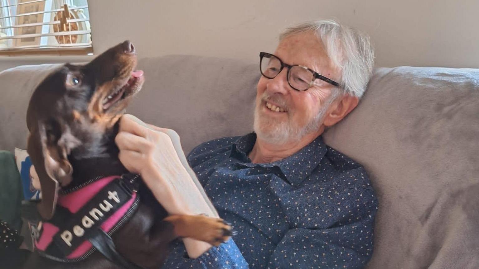 Tim Strugnell sitting on a sofa while wearing a shirt and glasses. There is a small brown dog on his lap which he is stroking. He is smiling and seems happy.