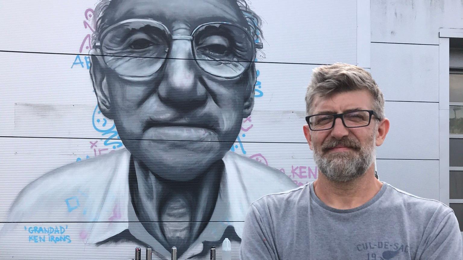 A mural of an elderly man, who has on glasses, looking straight on, painted on a building, with words behind the image and a signature at the bottom. A man is standing beside the image, with short hair, glasses and is hearing a grey T-shirt. 