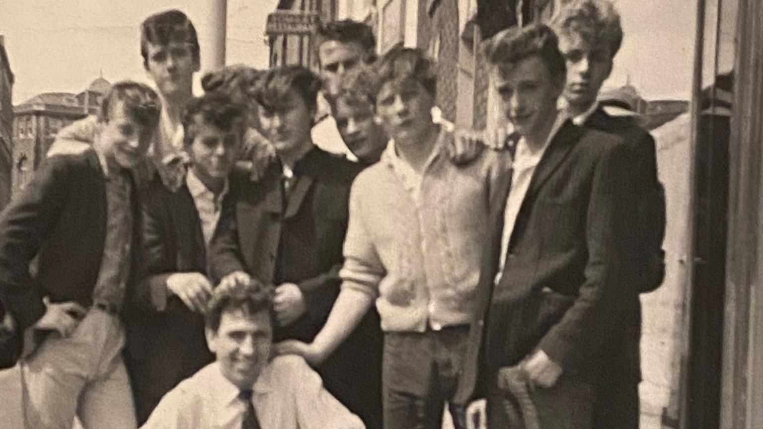 A black and white photo of lots of young men gathered round a squatting man in a white shirt and black tie in a town centre. 