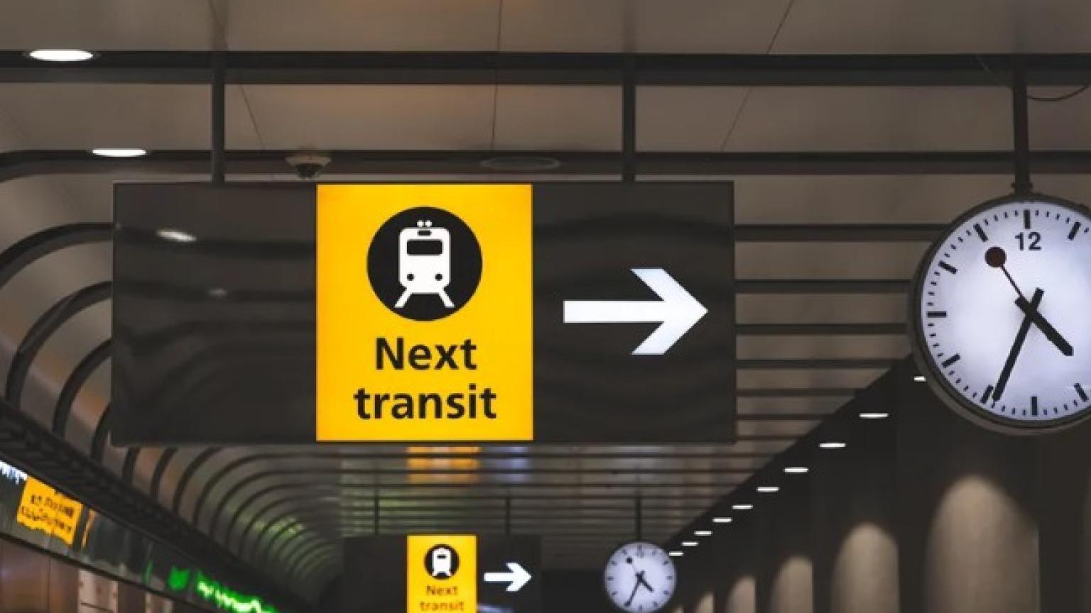 A yellow sign with the words "Next transit" written in black. The sign is hanging from the ceiling inside a tunnel. To the right is a white clock face showing the time 16:34.