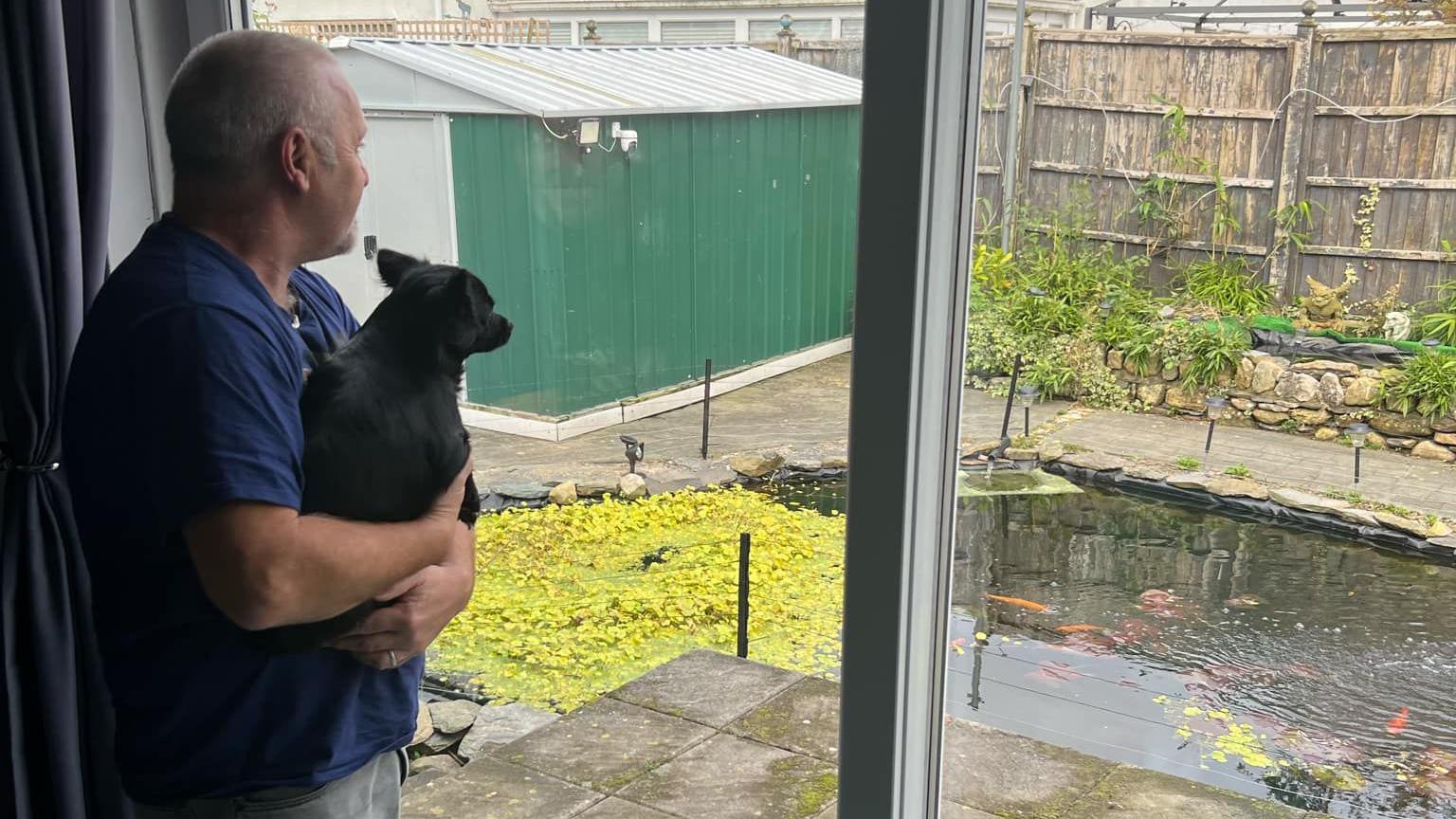 Nige Cooper, holding his dog in his hand, looks out to his pond, with lots of fish swimming in, from his window.