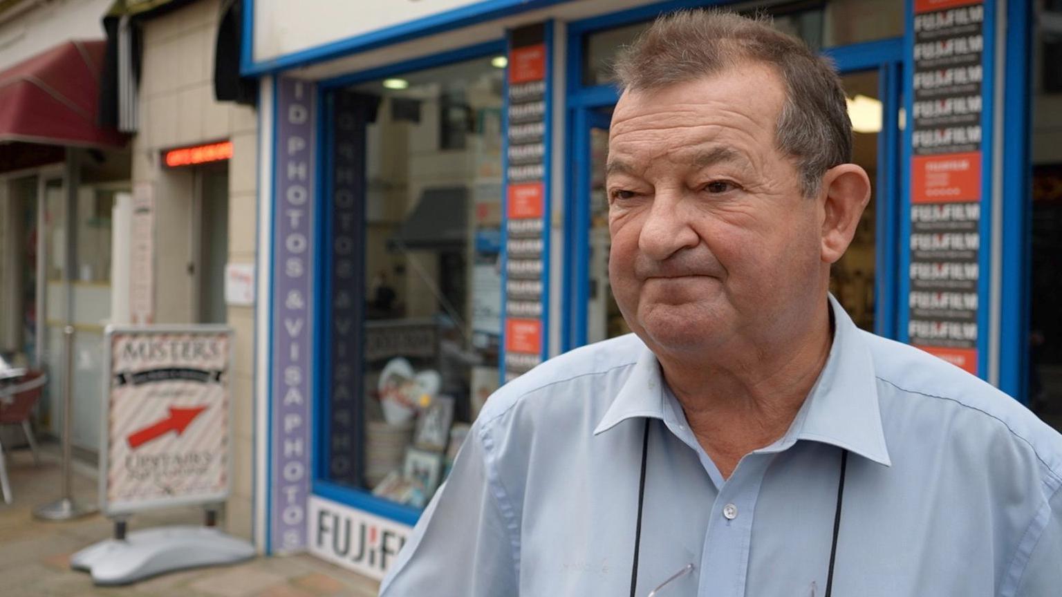 Mark Fisher is wearing a blue shirt, he is standing outside the Fotosound shop. 