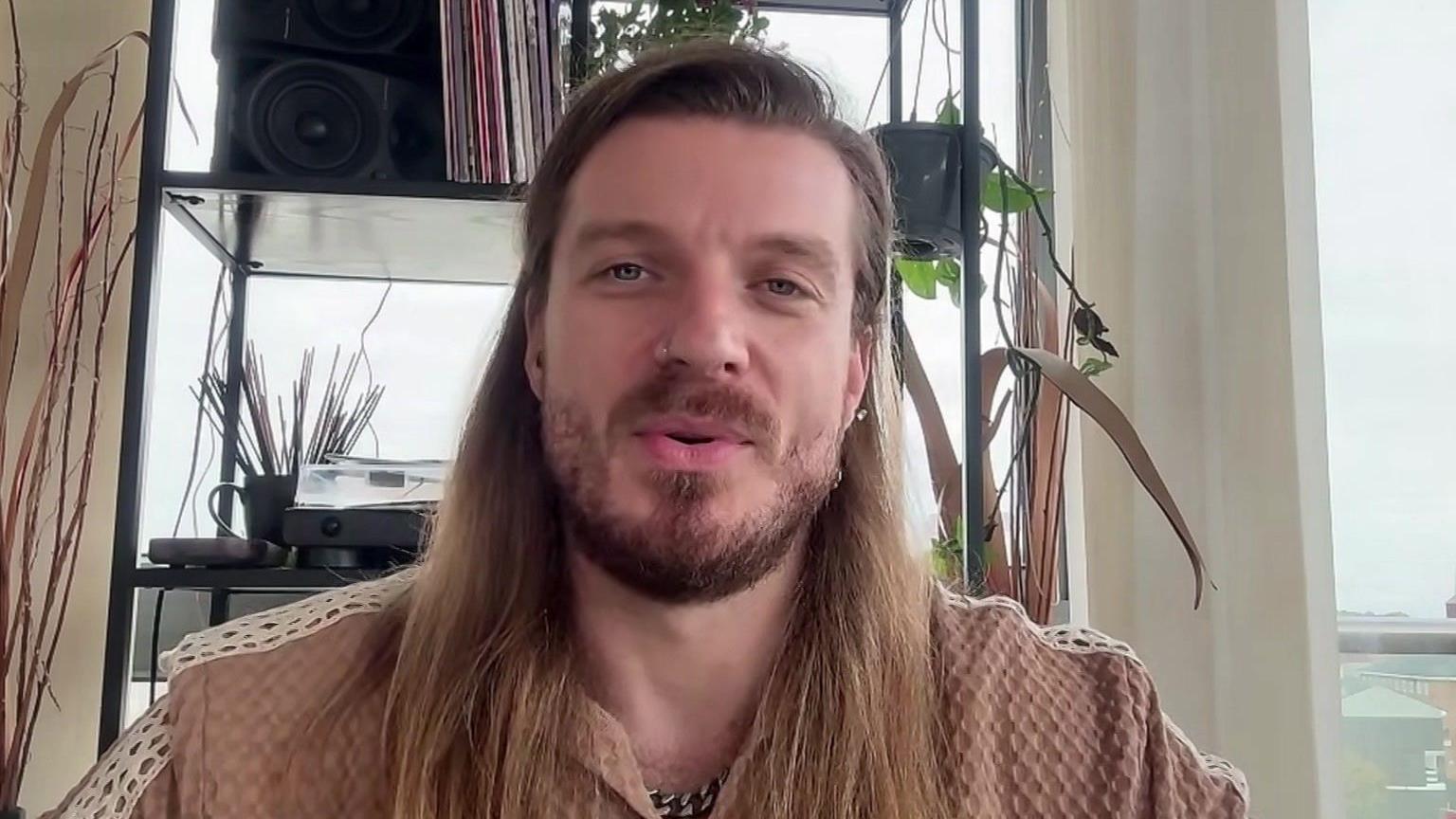 Dom Topics, who has long blond hair, sits in his lounge talking to the BBC