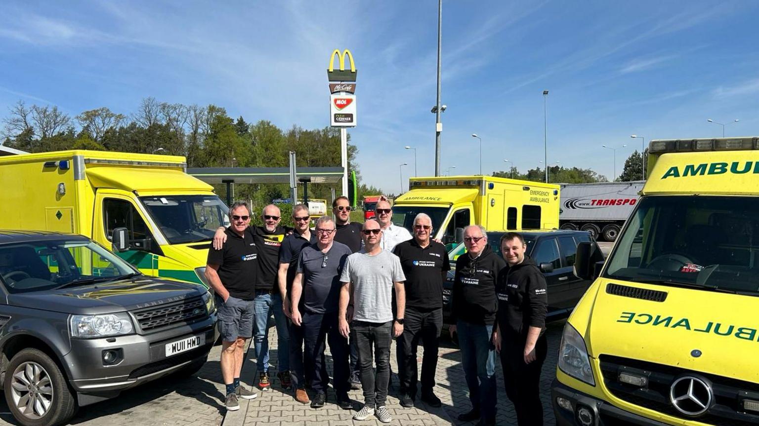 Ten men at a European service station with three old ambulances. Seven are wearing black shirts, two grey and one white. They also have two Land Rover vehicles. One is grey, one is black.