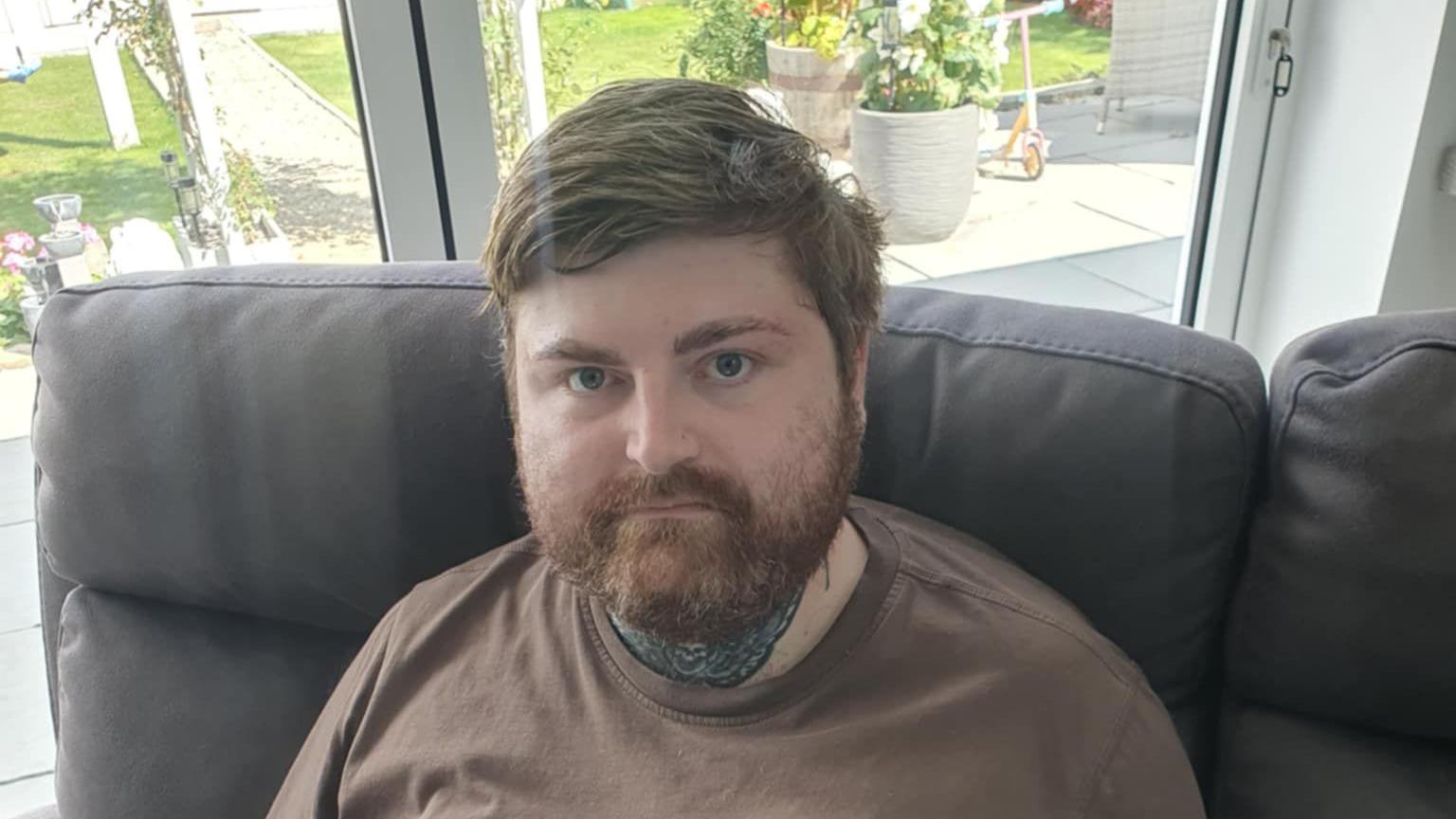 Mr Luccarini sits on a black leather sofa. He has brown hair and thick beard. He wears a brown T-shirt. Windows overlooking a garden can be seen behind him.