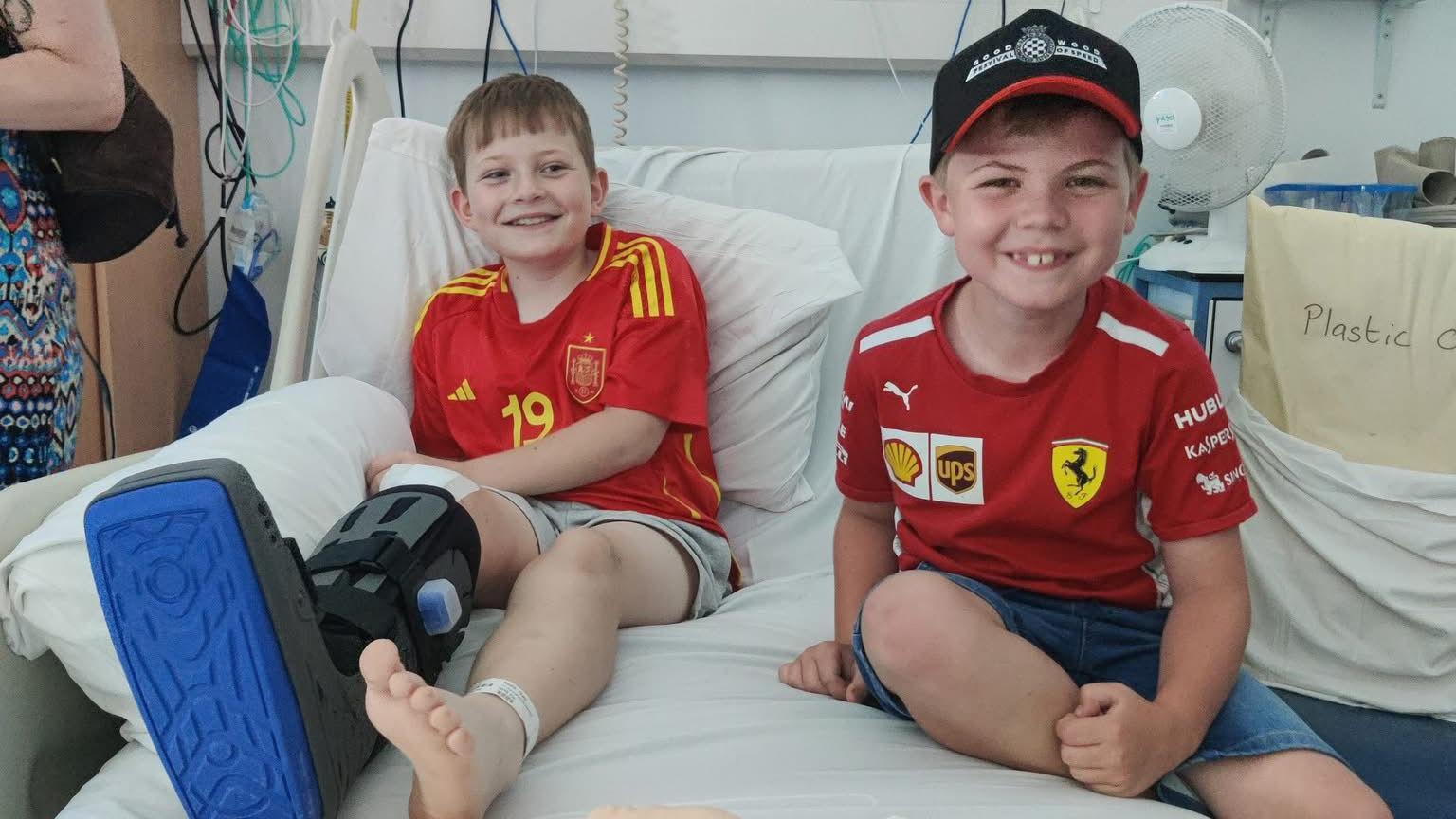 Theo Rees visited by his best friend Jacob, with both sitting on a hospital bed. Both are wearing red t-shirts and shorts and are smiling. Theo is wearing a big black and blue boot on one foot.